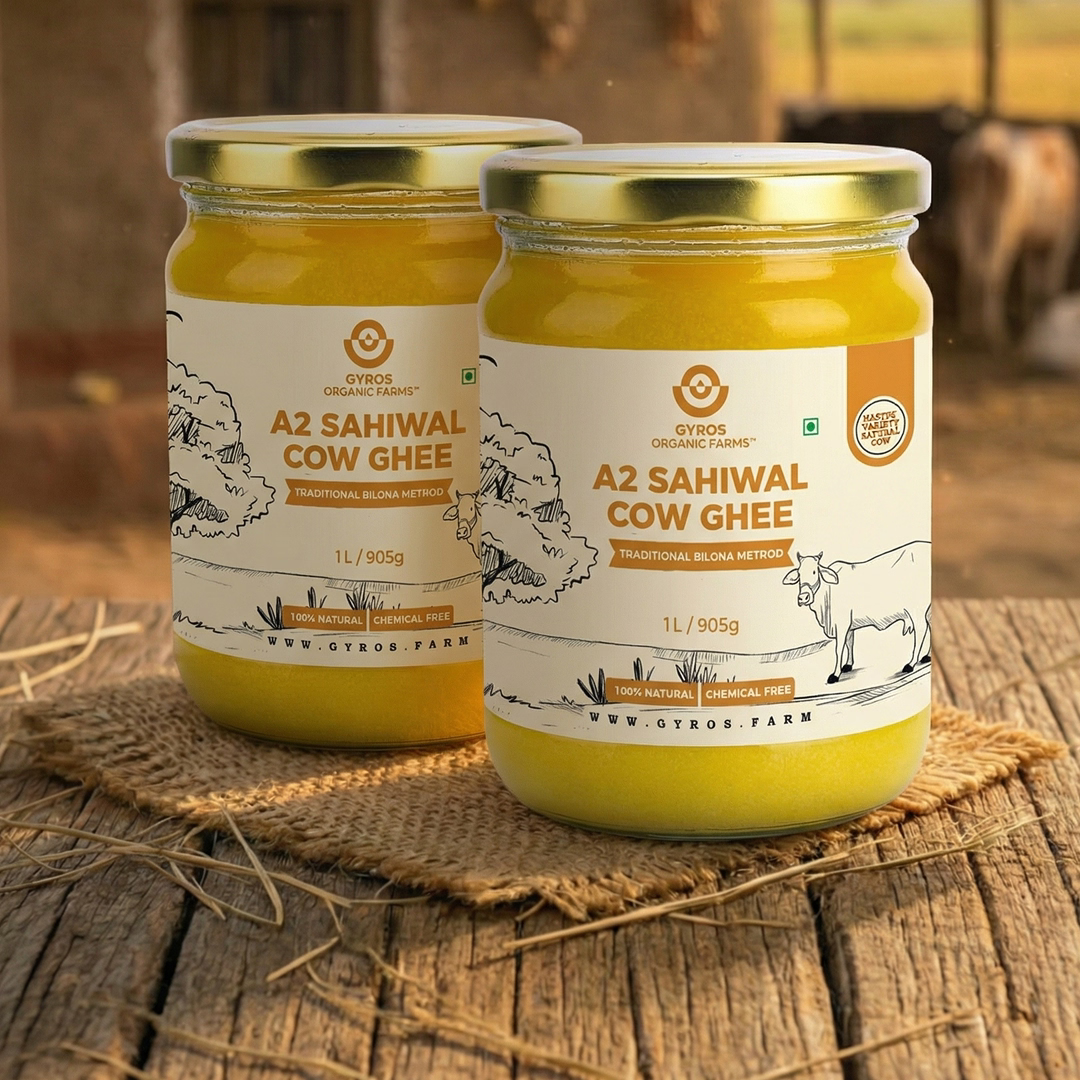 Two jars of Gyros A2 Sahiwal cow ghee placed on a rustic wooden surface with a village farm background.