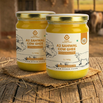 Two jars of Gyros A2 Sahiwal cow ghee placed on a rustic wooden surface with a village farm background.