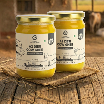 Two jars of Gyros Organic Farms A2 Desi Cow Ghee on a rustic wooden surface with a blurred farm background.